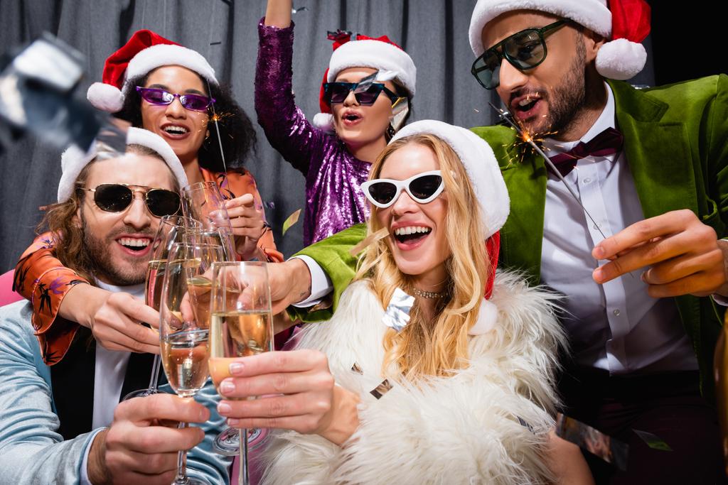 χαμογελαστοί διαφυλετικοί φίλοι με καπέλα Σάντα γιορτάζουν το νέο έτος κοντά γκρι κουρτίνα σε μαύρο φόντο - Φωτογραφία, εικόνα