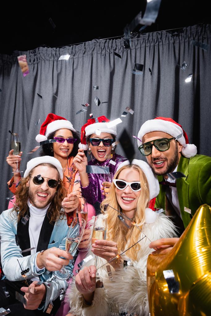 stilvolle interrassische Freunde in Weihnachtsmützen und Sonnenbrillen bei einer Party in der Nähe des grauen Vorhangs auf schwarzem Hintergrund - Foto, Bild