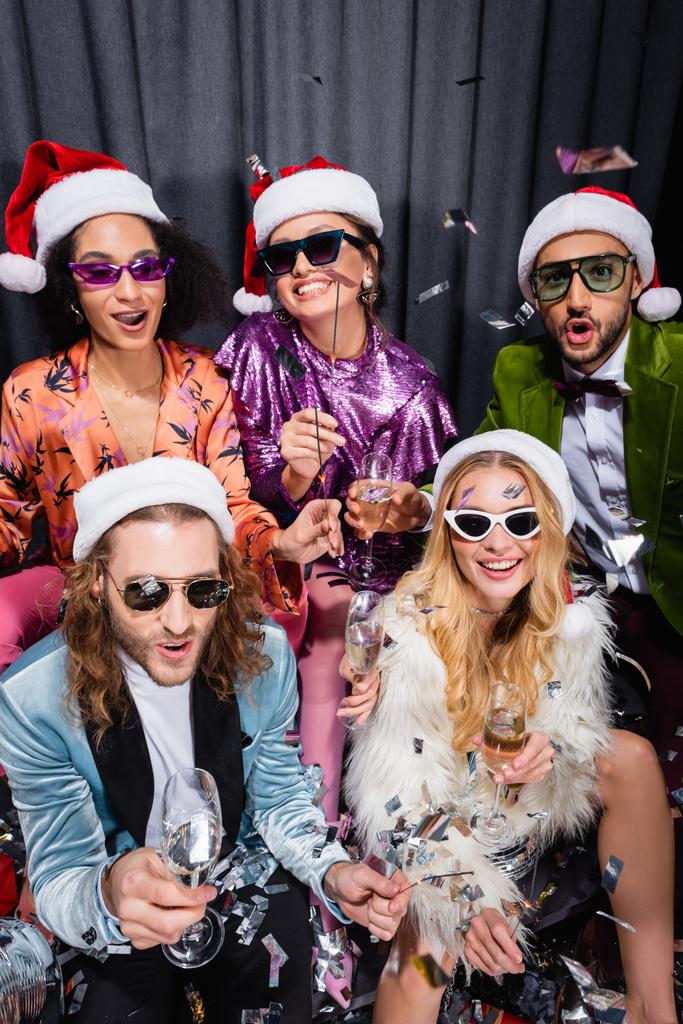 young adult interracial friends in santa hats and sunglasses having party on grey background - Photo, Image
