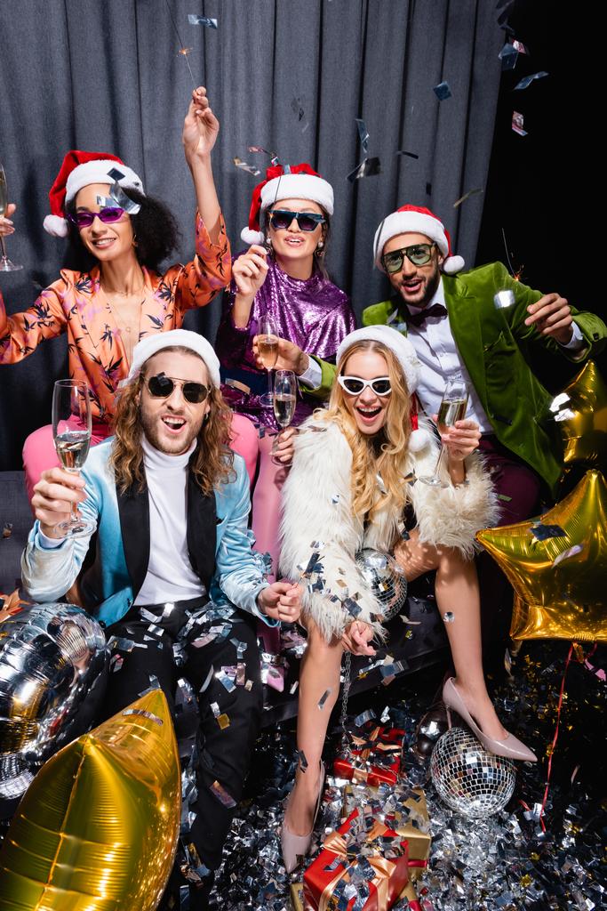 playful interracial friends in santa hats and sunglasses celebrating new year on grey couch on black background - Photo, Image