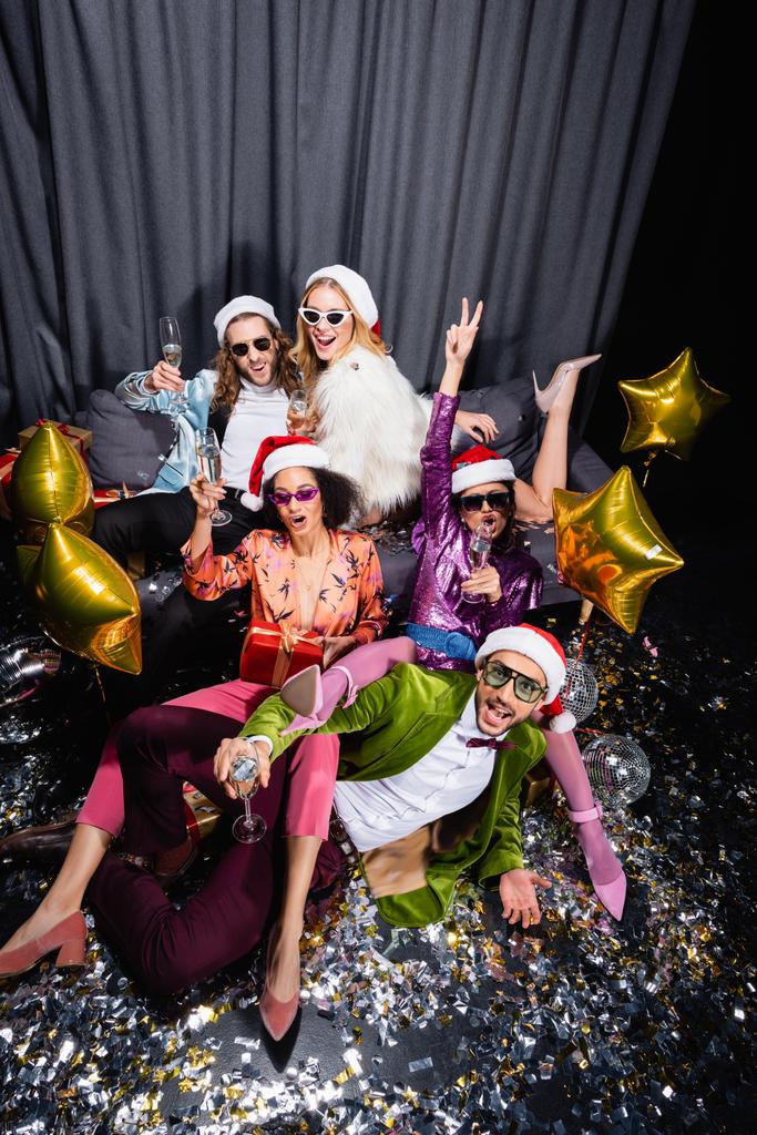παιχνιδιάρηδες διαφυλετικοί φίλοι με καπέλα santa γιορτάζουν το νέο έτος κοντά γκρι κουρτίνα σε μαύρο φόντο - Φωτογραφία, εικόνα