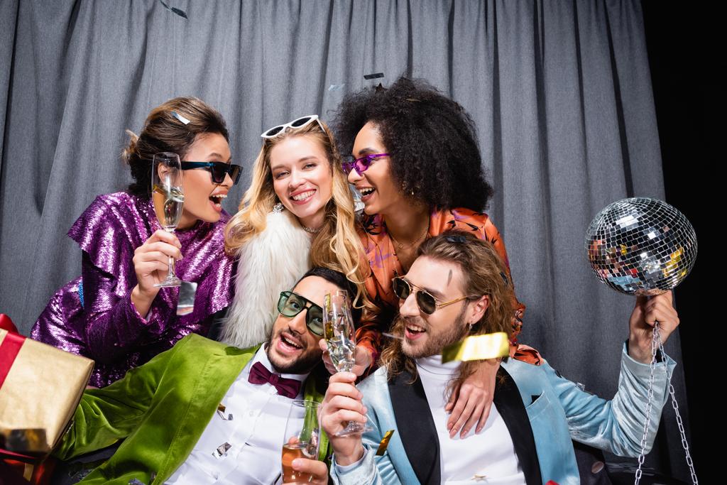 speelse interraciale vrienden in kleurrijke kleren drinken champagne in de buurt grijs gordijn op zwarte achtergrond - Foto, afbeelding