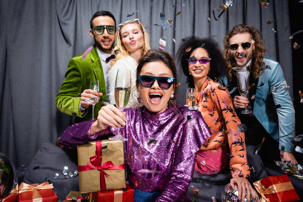 playful interracial friends with gift boxes drinking champagne on grey background - Photo, Image