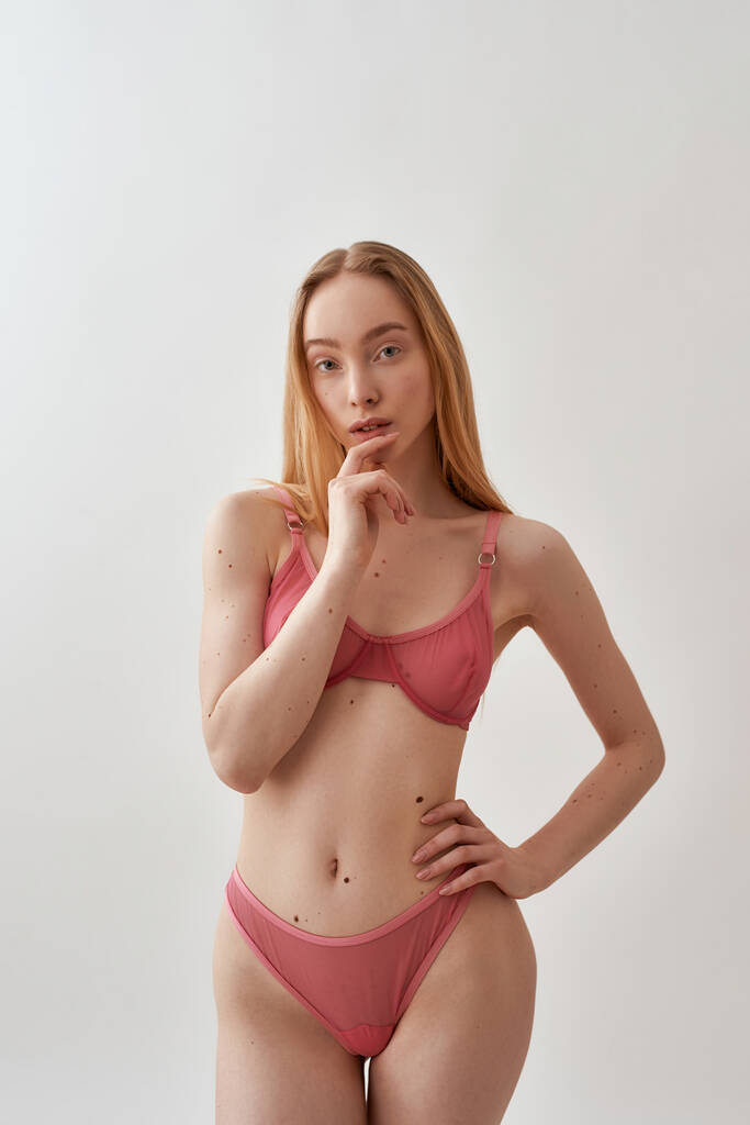 Χαριτωμένη νεαρή καυκάσια γυναίκα με λεπτό σώμα φορώντας ροζ διαφανές εσώρουχο κοιτάζοντας την κάμερα, κρατώντας το χέρι κοντά στα χείλη της, ενώ θέτουν απομονώνονται σε ανοιχτό γκρι φόντο - Φωτογραφία, εικόνα