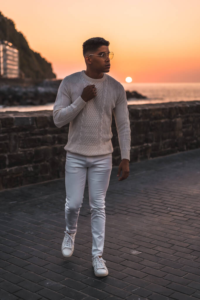 Καλοκαιρινός τρόπος ζωής, ένας νεαρός Λατίνος που απολαμβάνει τις καλοκαιρινές διακοπές. ντυμένοι στα λευκά με παντελόνι και πουλόβερ, περπατώντας δίπλα στη θάλασσα - Φωτογραφία, εικόνα