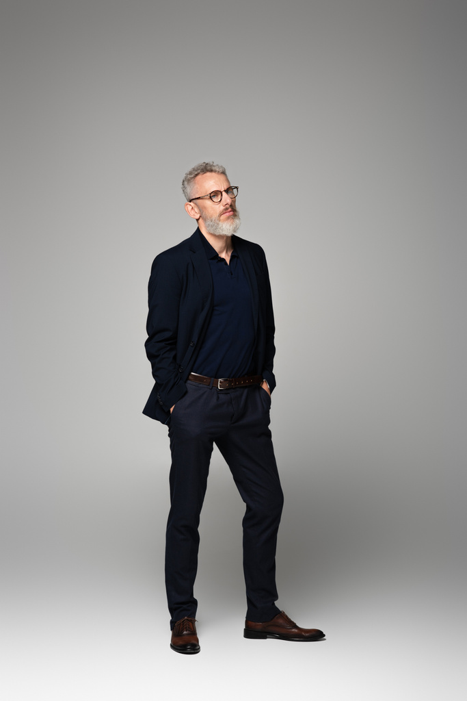 longitud completa del hombre de mediana edad en gafas y traje de pie con las manos en los bolsillos en gris - Foto, imagen