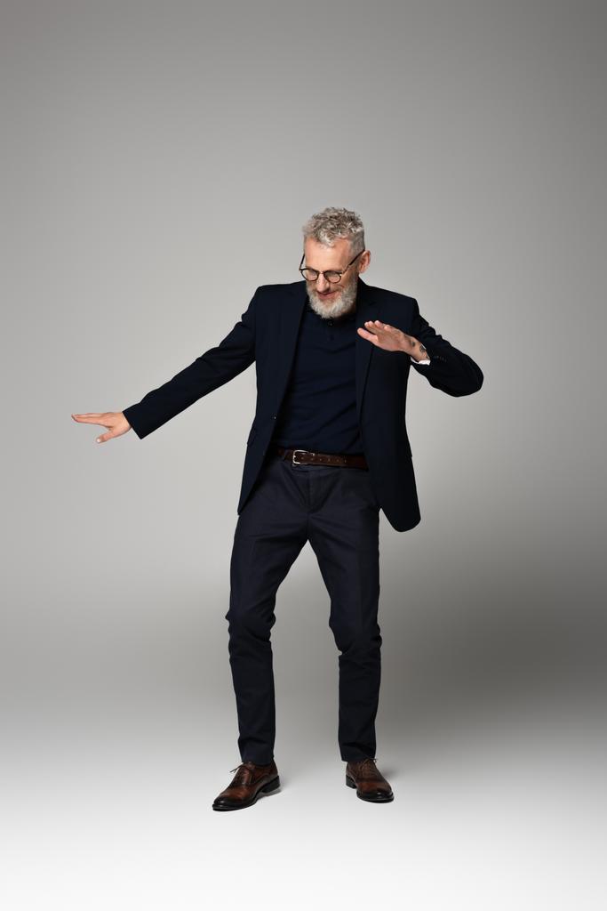 volledige lengte van vrolijke man van middelbare leeftijd in bril en pak dansen op grijs - Foto, afbeelding