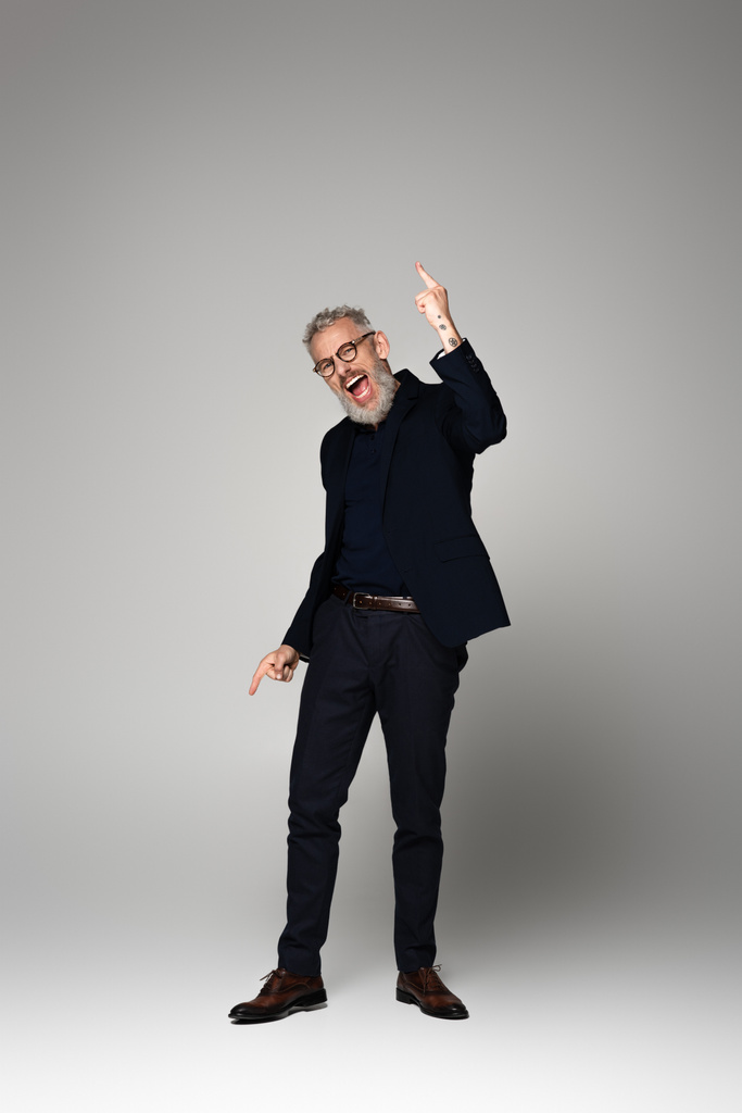 longitud completa del hombre de mediana edad excitado en gafas y traje apuntando con los dedos en gris - Foto, imagen