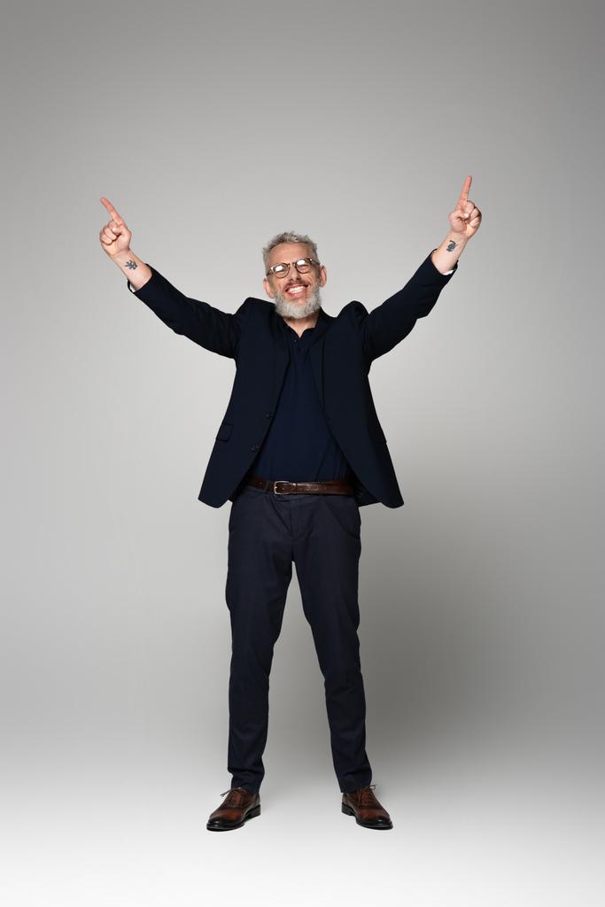 полная длина улыбающийся мужчина средних лет в очках и костюме указывая пальцами на серый - Фото, изображение