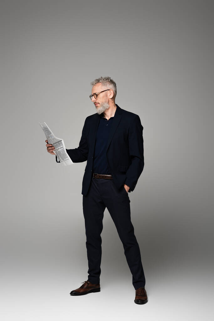 longitud completa del hombre de mediana edad en gafas y traje leyendo periódico mientras posando en gris - Foto, Imagen