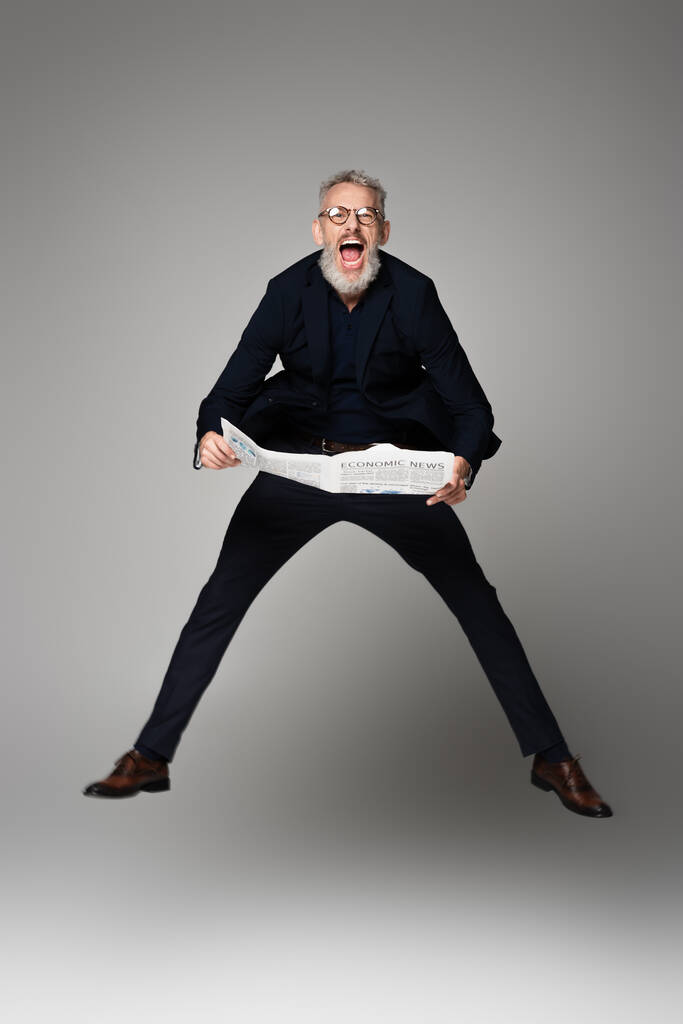 plná délka užaslého muže v brýlích a obleku držícího noviny při levitaci na šedé - Fotografie, Obrázek