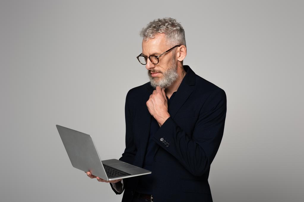 nachdenklicher Mann mittleren Alters mit grauen Haaren im Anzug mit Laptop isoliert auf grau - Foto, Bild