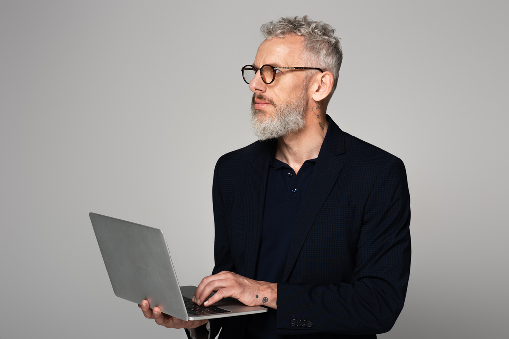 задумчивый мужчина средних лет с седыми волосами в костюме, держащий ноутбук изолирован на сером - Фото, изображение