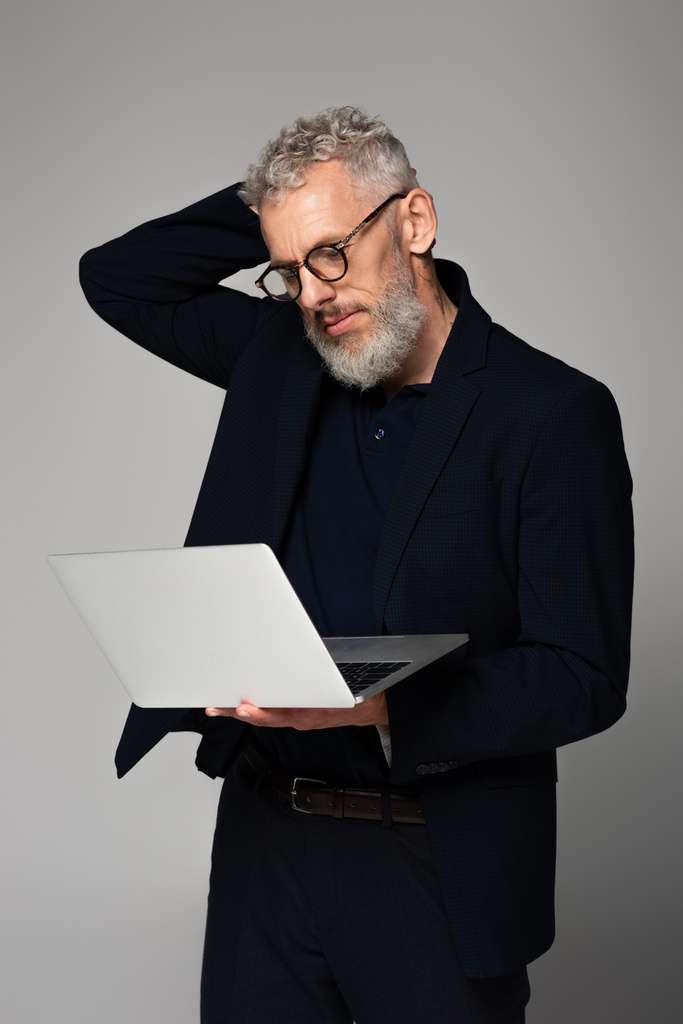 mietteliäs keski-ikäinen mies silmälaseissa katselemassa harmaalla eristettyä kannettavaa tietokonetta - Valokuva, kuva