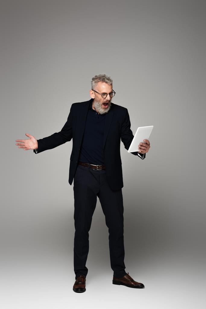 longitud completa de hombre de negocios enojado de mediana edad con el pelo gris gritando mientras mira la tableta digital en gris - Foto, imagen