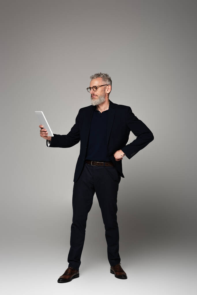 volledige lengte van de middelbare leeftijd zakenman met grijs haar kijken naar digitale tablet en poseren op grijs - Foto, afbeelding