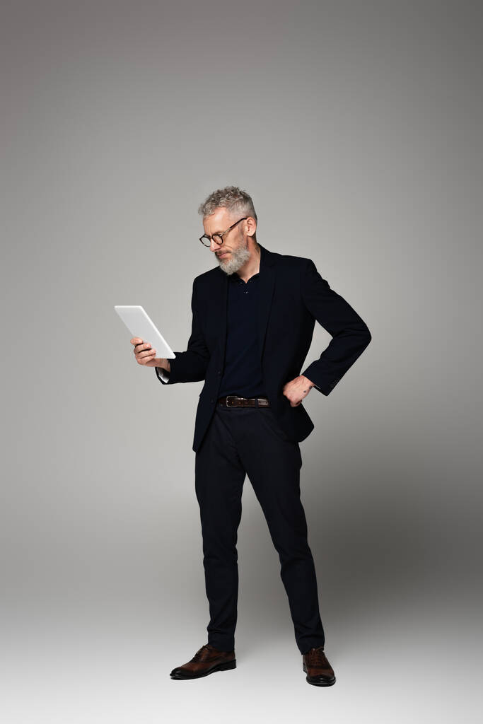 Mann mit grauen Haaren blickt auf digitales Tablet und posiert auf grau - Foto, Bild