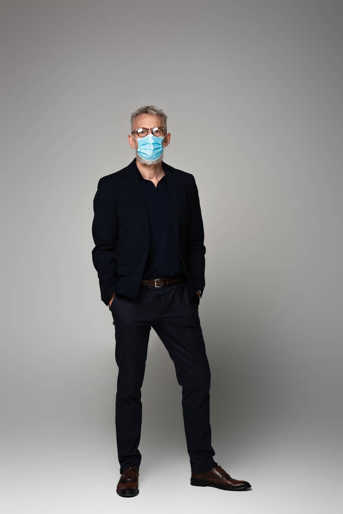 longitud completa de hombre maduro con pelo gris en gafas y máscara médica posando con las manos en bolsillos en gris  - Foto, Imagen