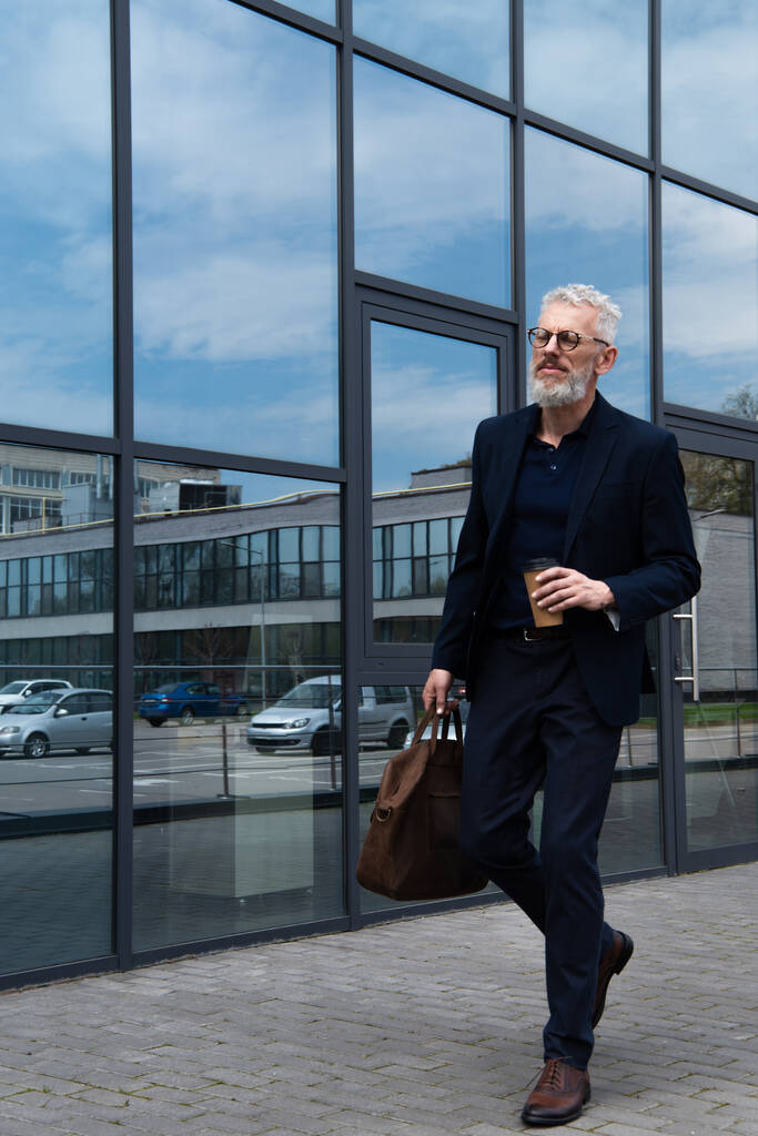 comprimento total do homem maduro com cabelos grisalhos segurando saco de couro e copo de papel enquanto caminhava perto do edifício moderno  - Foto, Imagem