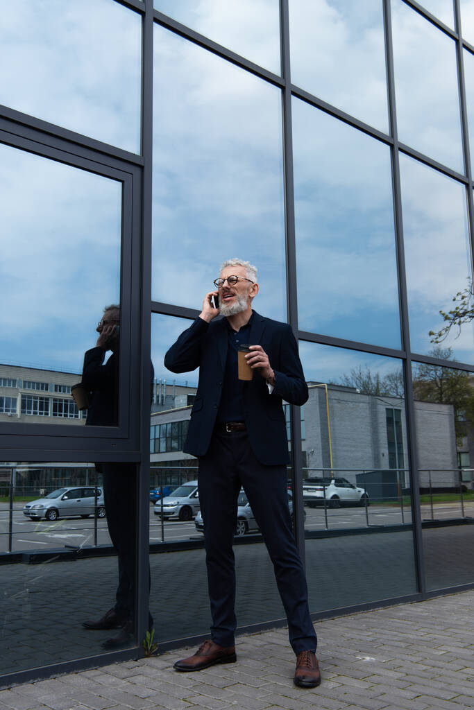 повнометражний зрілий чоловік з сірим волоссям тримає паперову чашку під час розмови на смартфоні біля сучасної будівлі
  - Фото, зображення