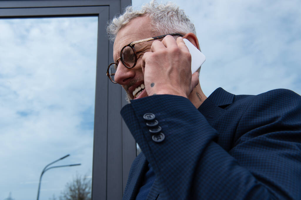 šťastný podnikatel s šedými vlasy mluví na smartphone v blízkosti moderní budovy  - Fotografie, Obrázek