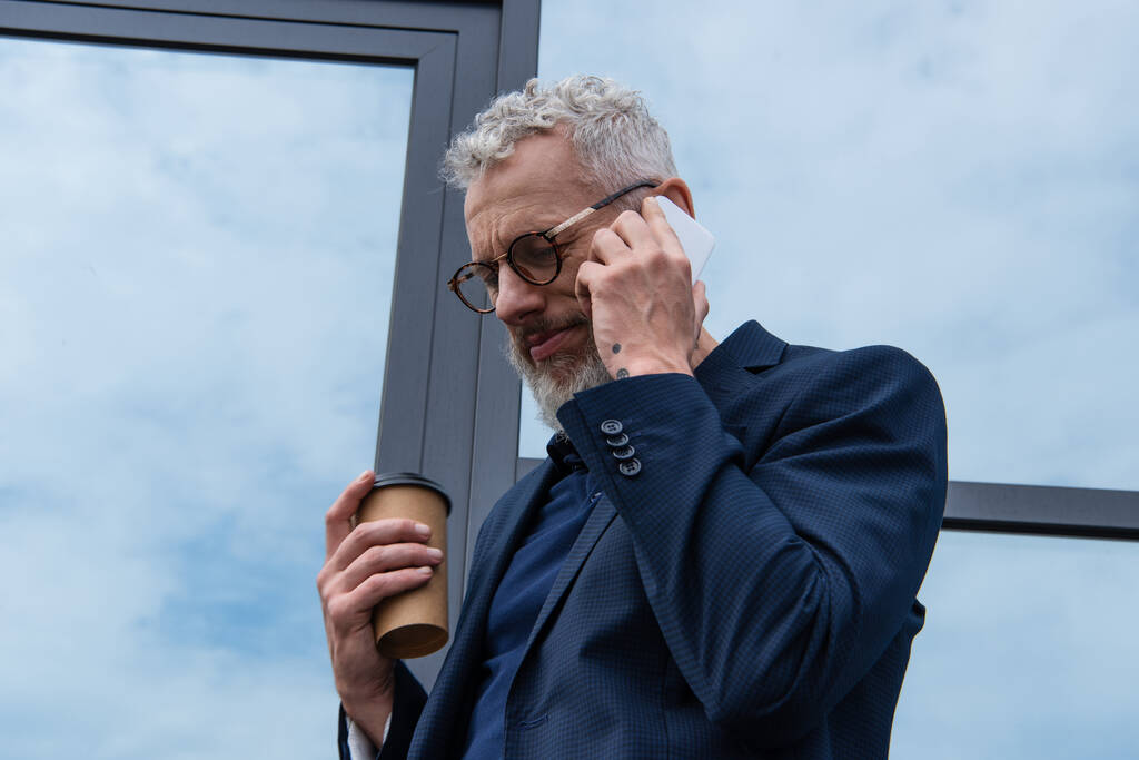 мужчина с седыми волосами в очках держит бумажную чашку во время разговора на смартфоне возле современного здания  - Фото, изображение