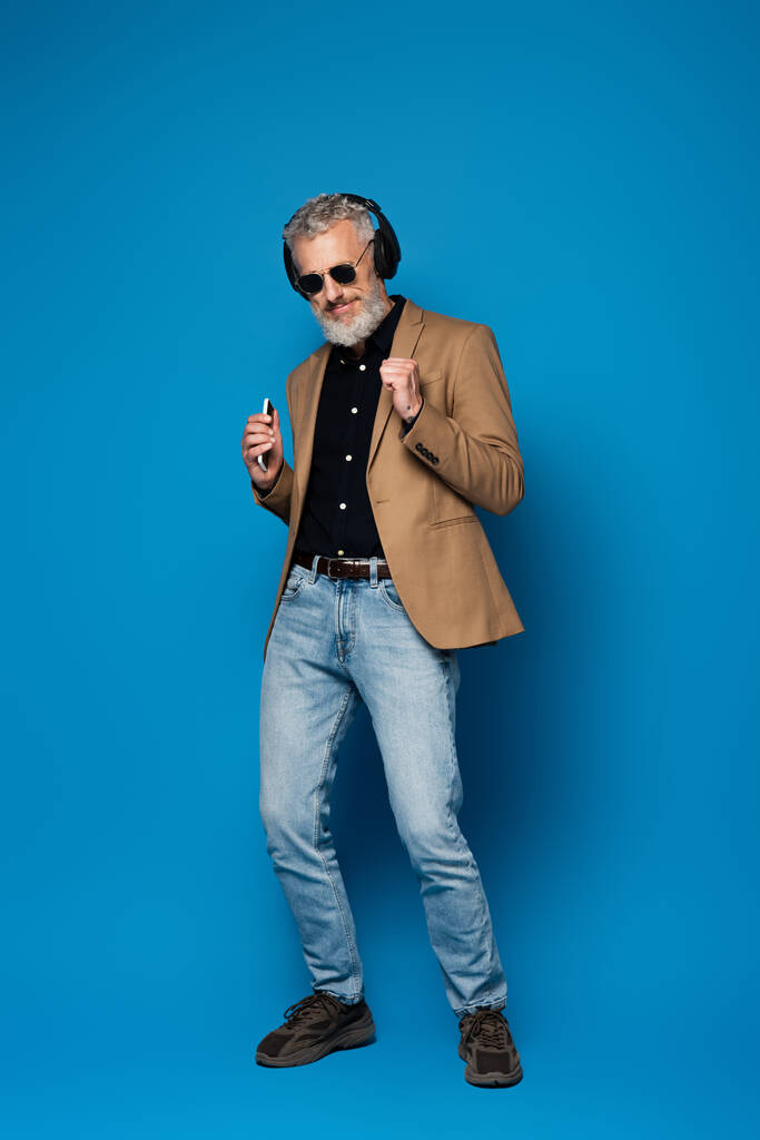 полный рост довольного мужчины средних лет в солнцезащитных очках, держащего смартфон и слушающего музыку в беспроводных наушниках синего цвета - Фото, изображение