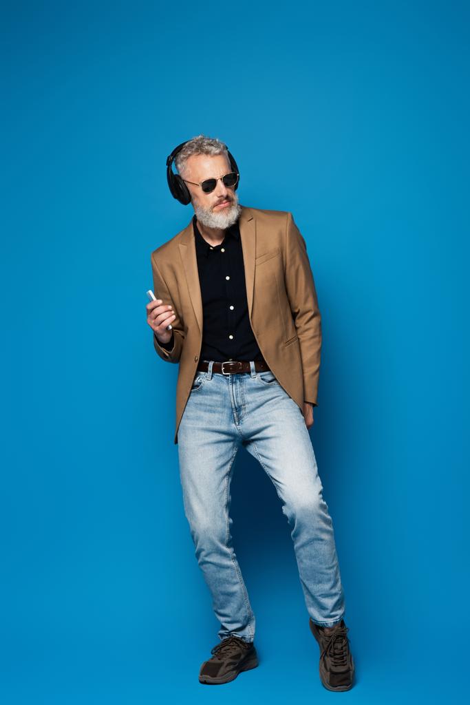 Мужчина средних лет в солнцезащитных очках в полный рост держит смартфон и слушает музыку в наушниках синего цвета - Фото, изображение