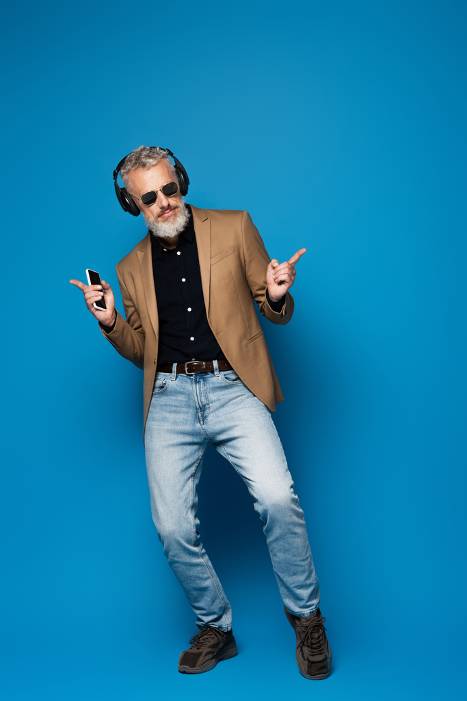 pełna długość uśmiechniętego mężczyzny w średnim wieku w okularach przeciwsłonecznych trzymającego smartfona podczas słuchania muzyki w słuchawkach na niebiesko - Zdjęcie, obraz