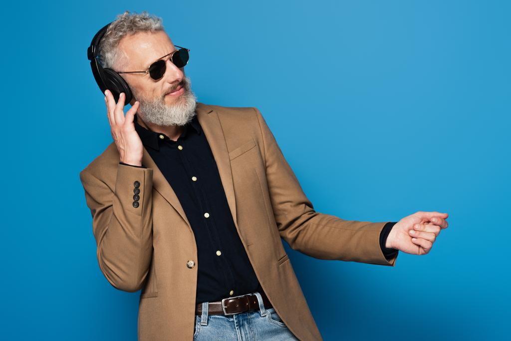 задоволений чоловік середнього віку в сонцезахисних окулярах слухає музику в навушниках і танцює на синьому
 - Фото, зображення