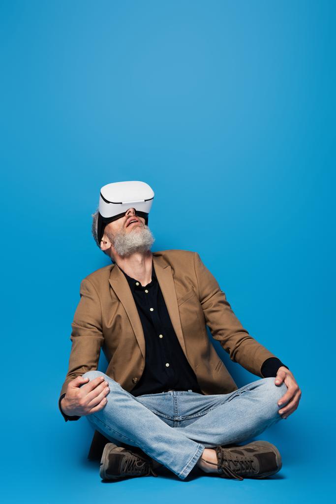 volledige lengte van de man van middelbare leeftijd in vr headset zitten met gekruiste benen en kijken omhoog op blauw  - Foto, afbeelding