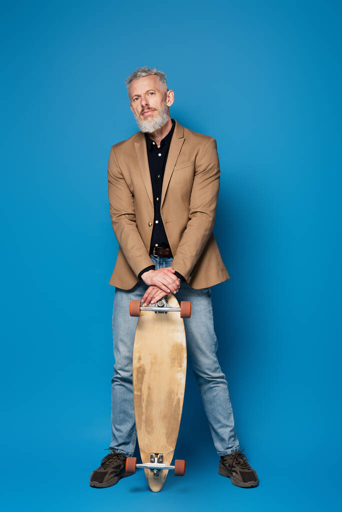 Полная длина бородатого мужчины средних лет, стоящего с лонгбордом на голубом фоне  - Фото, изображение