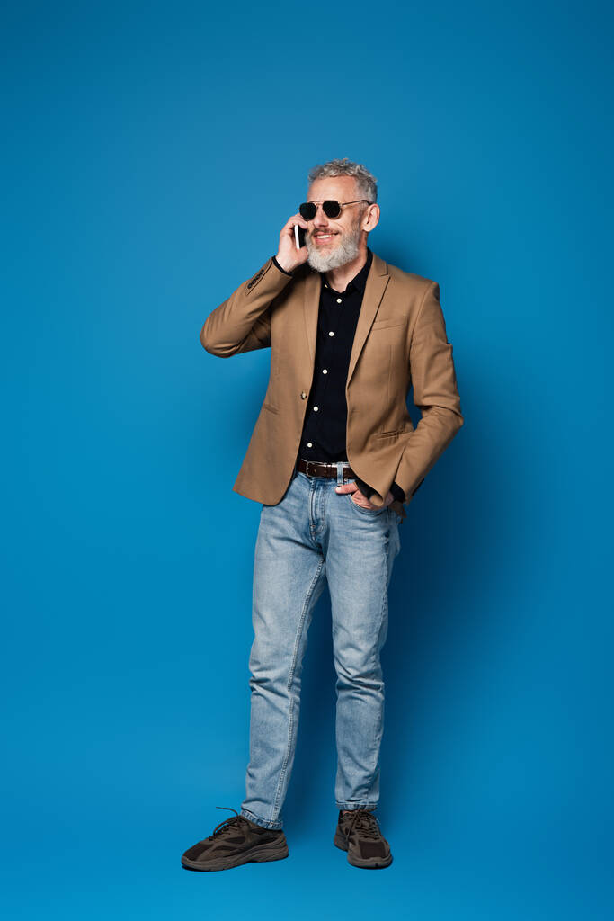 volledige lengte van gelukkig middelbare leeftijd man in zonnebril praten op mobiele telefoon op blauw - Foto, afbeelding
