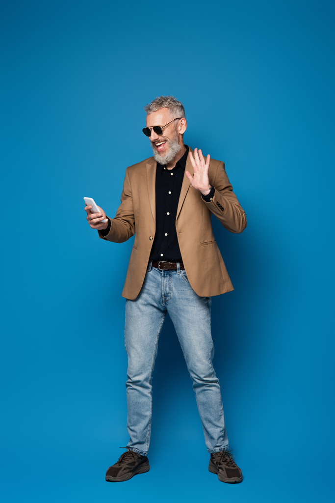 in voller Länge glücklicher Mann mittleren Alters mit Sonnenbrille, Videoanruf auf dem Handy und winkender Hand auf blauem Grund - Foto, Bild