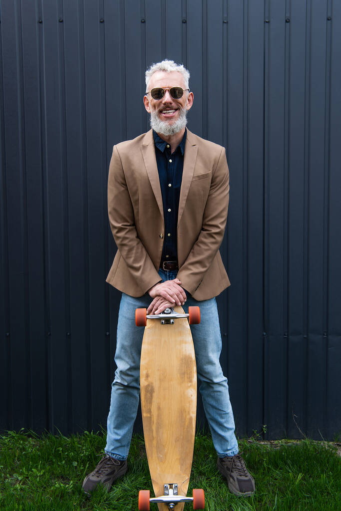 volledige lengte van gelukkig man van middelbare leeftijd in zonnebril houden longboard buiten  - Foto, afbeelding