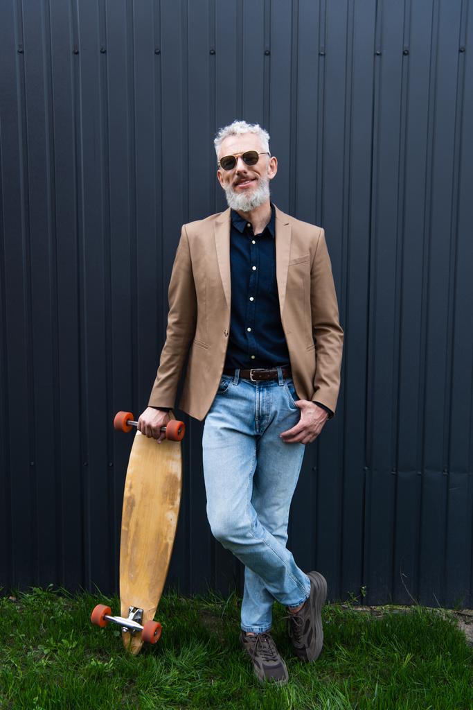 volledige lengte van vrolijke man van middelbare leeftijd in zonnebril houden longboard buiten  - Foto, afbeelding