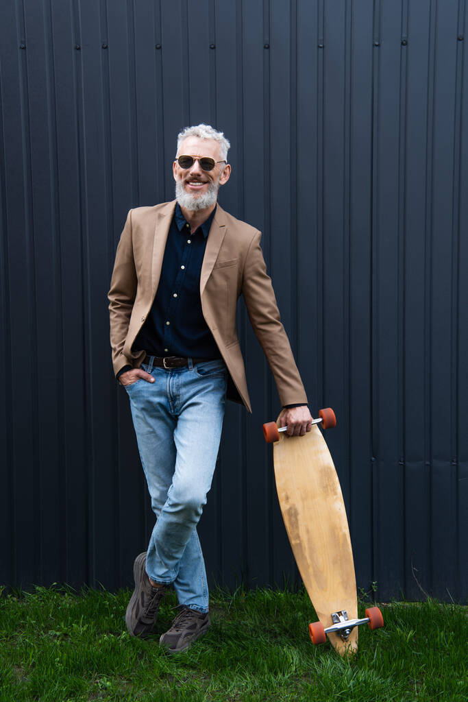 volledige lengte van tevreden man van middelbare leeftijd in zonnebril houden longboard buiten  - Foto, afbeelding