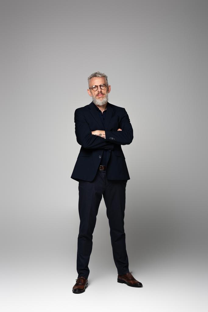 longitud completa de hombre de negocios en gafas y traje posando con los brazos cruzados en gris - Foto, imagen