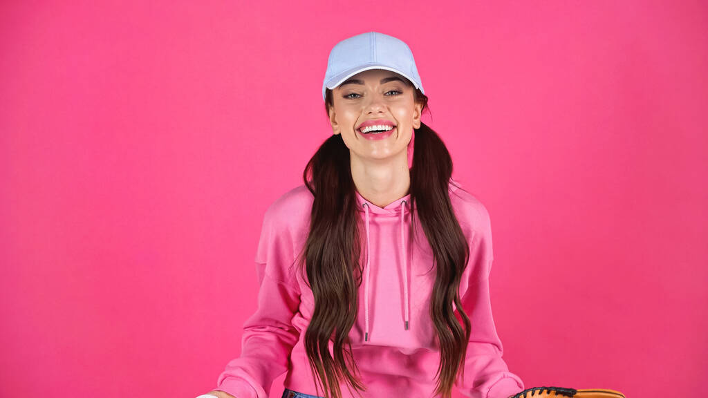 smiling young adult woman in cap and hoodie looking at camera isolated on pink - Photo, image