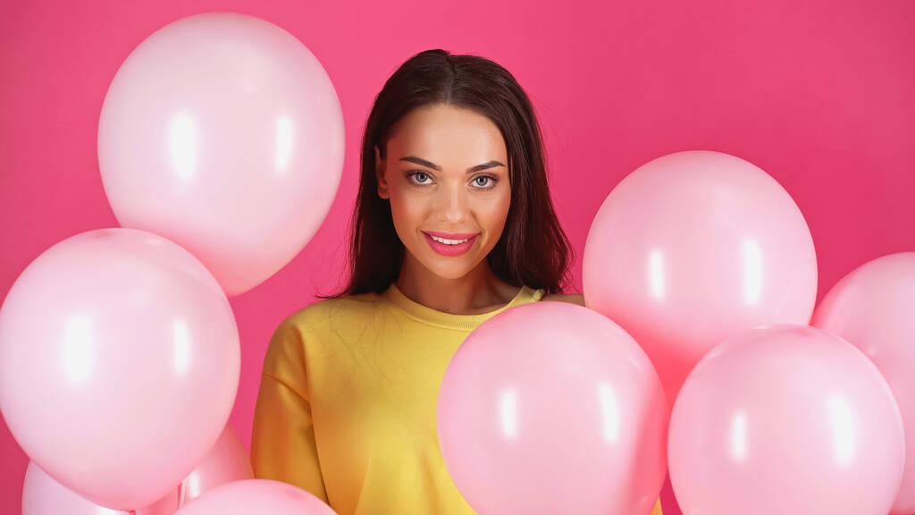Lächelnde junge erwachsene Frau in gelber Bluse zwischen Luftballons auf rosa - Foto, Bild