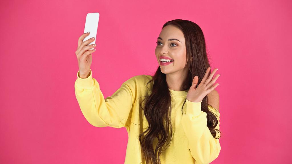 улыбающаяся молодая женщина с видеозвонком и размахивая рукой на мобильный телефон изолированы на розовый - Фото, изображение