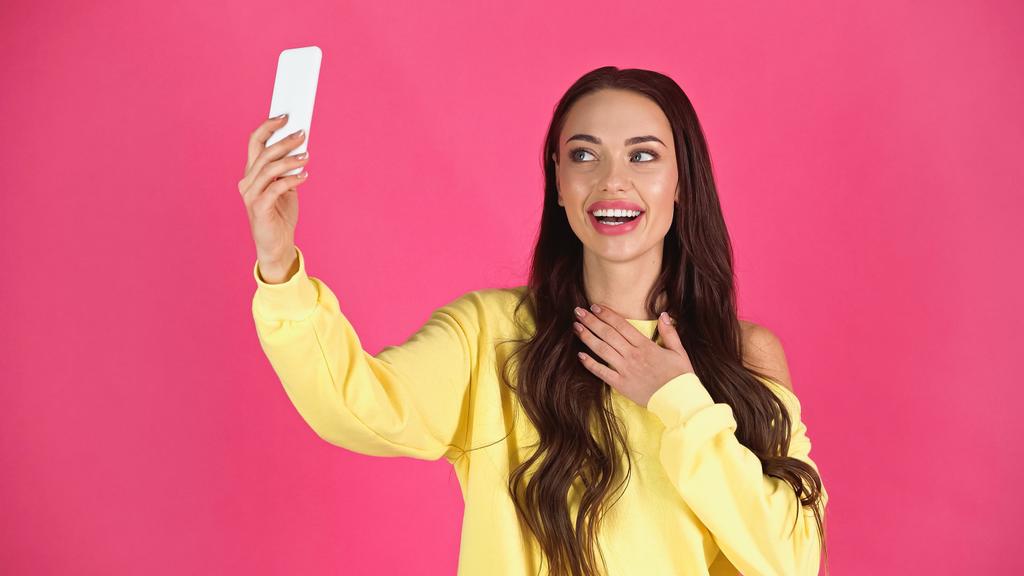 smiling young adult woman taking selfie on cellphone and holding hand on chest isolated on pink - Photo, image