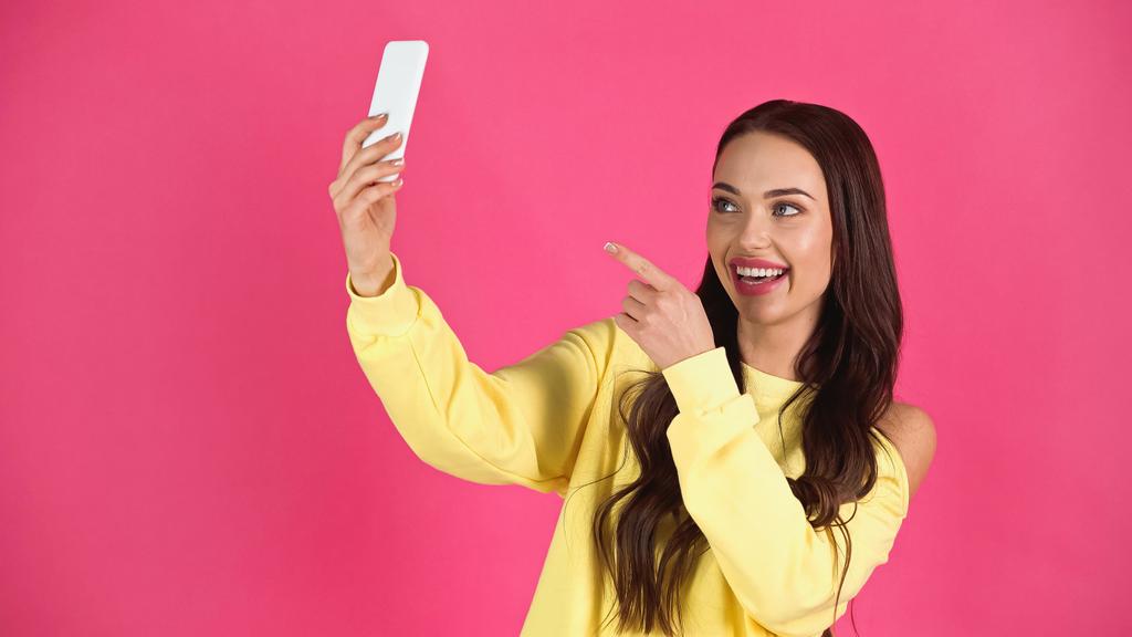 усміхнена молода доросла жінка бере селфі і вказує пальцем на мобільний телефон ізольовано на рожевому
 - Фото, зображення