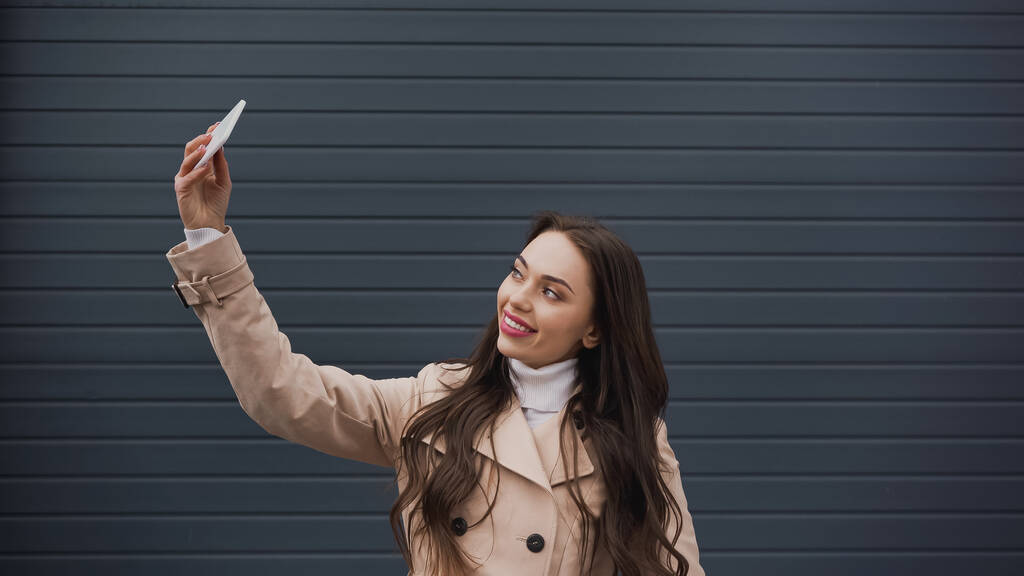 smiling young adult woman taking selfie with cellphone on textured grey background - Fotoğraf, Görsel