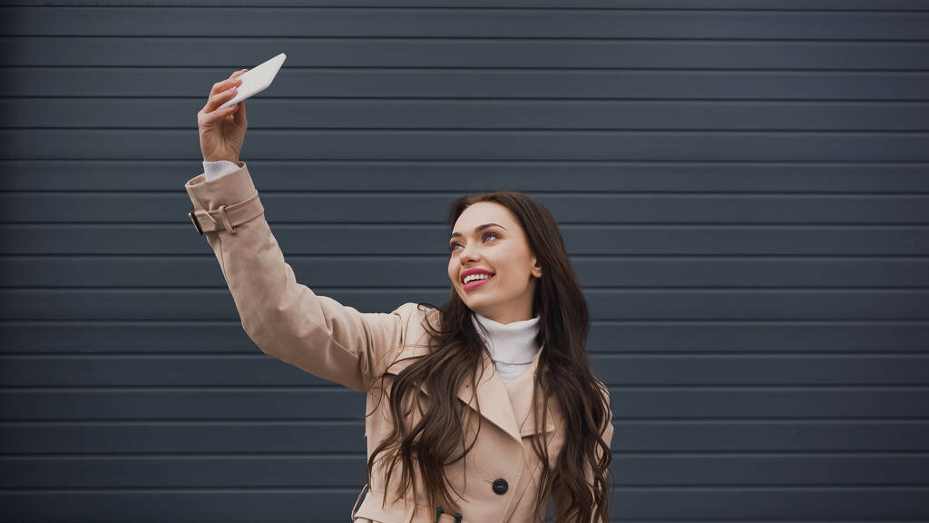 улыбающаяся молодая женщина в бежевом плаще делает селфи с сотовым телефоном на сером текстурированном фоне  - Фото, изображение