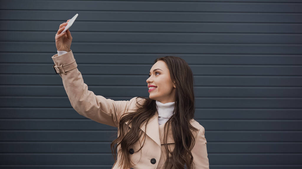 hübsche junge erwachsene Frau macht Selfie mit Handy auf grau strukturiertem Hintergrund - Foto, Bild