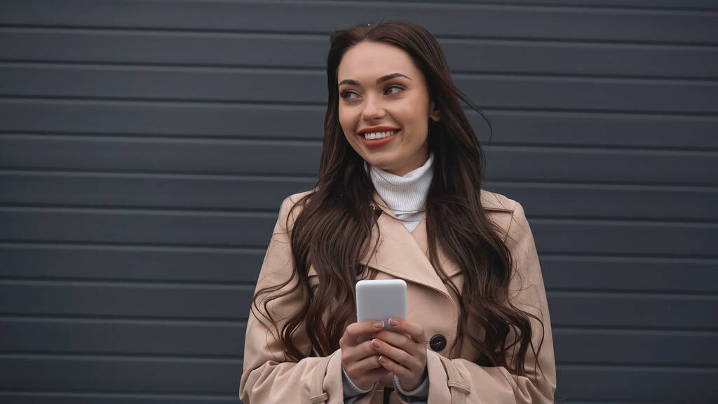 усміхнена молода доросла жінка тримає мобільний телефон в руках на сірому текстурованому фоні
 - Фото, зображення