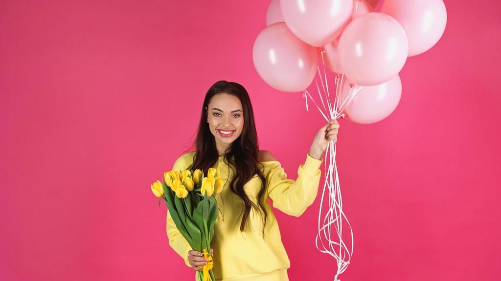 усміхнена молода доросла жінка тримає букет тюльпанів і повітряних кульок ізольовано на рожевому
 - Фото, зображення