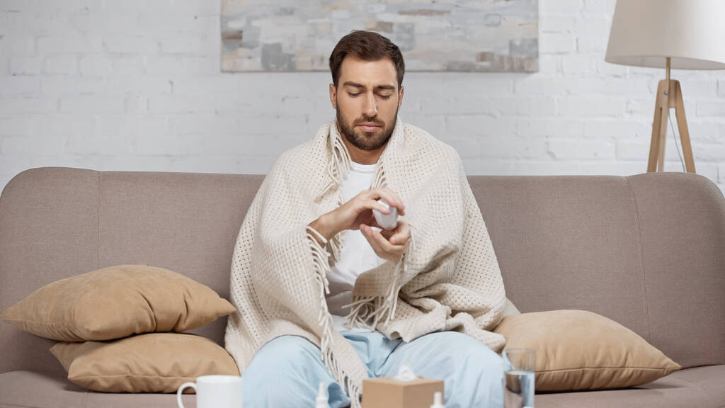 sick man sitting on sofa and holding bottle with pills near coffee table with drinks  - Photo, image