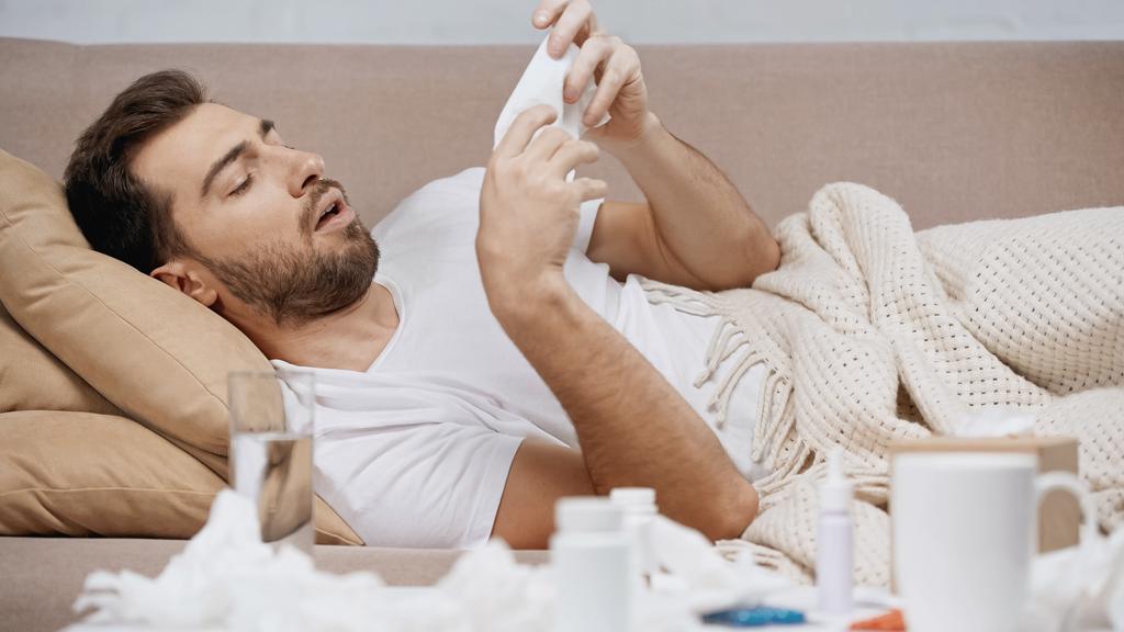 sick man lying under blanket and sneezing near medication and crumpled tissues  - Fotografie, Obrázek